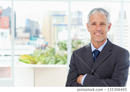 Middle-aged male standing with crossed arms in office by window panes with planter, copy space 135306405