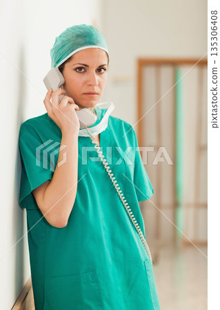 Female surgeon wearing green scrubs talking on corded telephone in hospital corridor, copy space 135306408