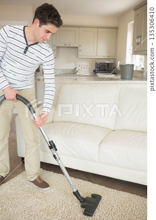 Man vacuuming shag rug near leather sofa and microwave-equipped kitchen with plant, copy space 135306410
