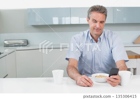 Middle-aged man holding smartphone and eating cereal at kitchen counter near white mugs and toaster Middle-aged man holding smartphone and eating cereal at kitchen counter near white mugs and toaster 135306415