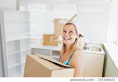 Woman holding large moving box with packing material by shelving in kitchen, unpacking new home 135306421