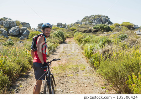 Male cyclist pausing beside mountain bike wearing helmet on dirt trail through rocky scrubland Male cyclist pausing beside mountain bike wearing helmet on dirt trail through rocky scrubland 135306424