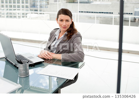 Businesswoman in gray blazer typing on laptop while reviewing documents at glass-top desk in office 135306426