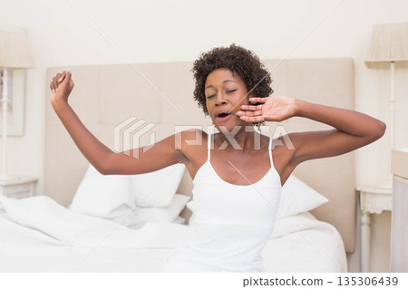 African American woman yawning and stretching on bed in bedroom with white linens and lamps 135306439