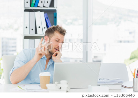 Male office worker leaning forward and pointing as using laptop and phone at desk Male office worker leaning forward and pointing as using laptop and phone at desk 135306440