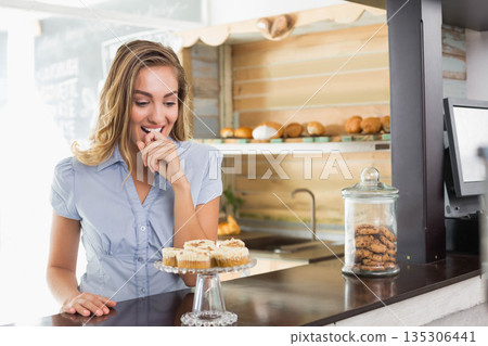 Woman in twenties gazing at glass cake stand with cupcakes, cookie jar at bakery, copy space 135306441