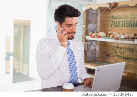 Business professional holding smartphone to ear and checking laptop at cafe with coffee cup Business professional holding smartphone to ear and checking laptop at cafe with coffee cup 135306453