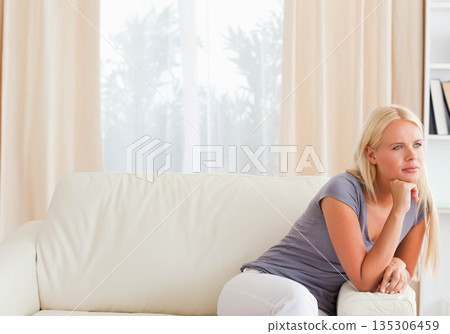 Woman in her twenties sitting on sofa in living room, leaning chin on fist, copy space 135306459