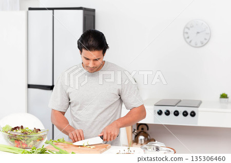 Mid adult male standing in white kitchen slicing celery on wooden cutting board beside glass bowl 135306460