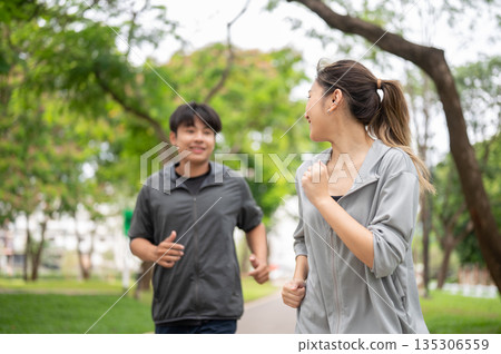 Asian woman and a man in sportswear talking together while exercising by running on pavement in park Asian woman and a man in sportswear talking together while exercising by running on pavement in park 135306559
