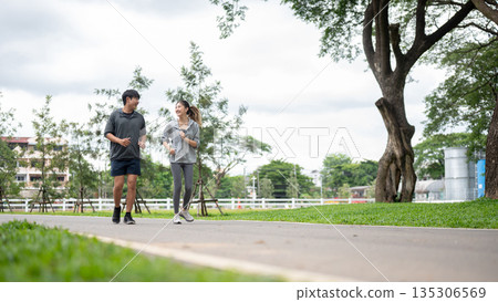 Asian woman and man in sportswear smiling and talking while running or jogging on pavement in park. 135306569