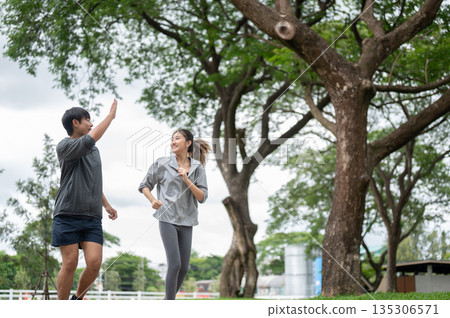 Asian man raising a hand and woman in sportswear smiling during a run or jogging on pavement in park Asian man raising a hand and woman in sportswear smiling during a run or jogging on pavement in park 135306571
