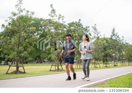 Asian woman in sportswear smiling talking during running or jogging with a man on pavement in park. Asian woman in sportswear smiling talking during running or jogging with a man on pavement in park. 135306584