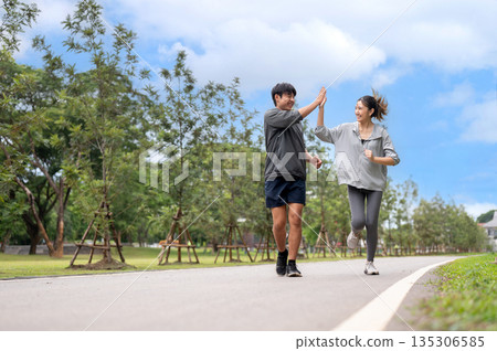 Asian woman in sportswear clapping or high five with a man during a run or jogging on pavement in park. 135306585