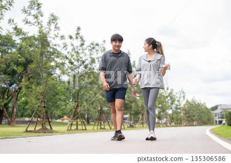 Asian woman in sportswear looking and talking with a man while walking together on pavement in park. 135306586