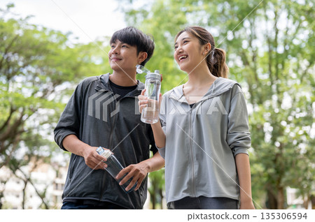 Asian man and woman in sportswear holding water bottle and looking same direction at park courtyard. 135306594