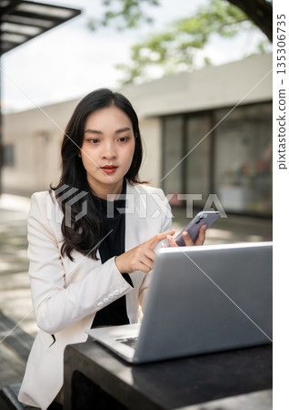 Asian female office worker businesswoman holding phone looking at laptop on table in cafe or garden. 135306735