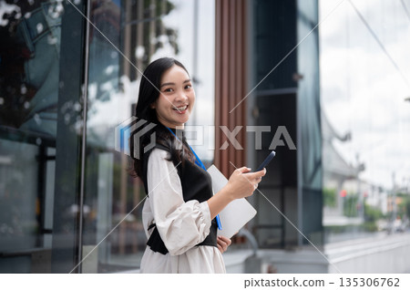 Smiling office worker or event staff holding folder looking away from phone standing out of building 135306762