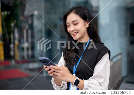 Smiling office worker or event staff holding and looking at phone while standing outside a building. Smiling office worker or event staff holding and looking at phone while standing outside a building. 135306763