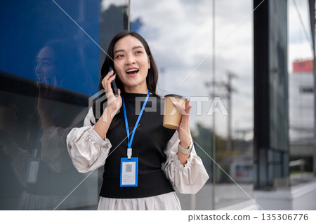 Smiling office worker or event staff holding coffee talking on phone while standing outside building 135306776
