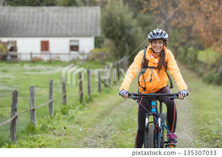 Female cyclist riding mountain bike in orange jacket along rural dirt path near fence, copy space Female cyclist riding mountain bike in orange jacket along rural dirt path near fence, copy space 135307033