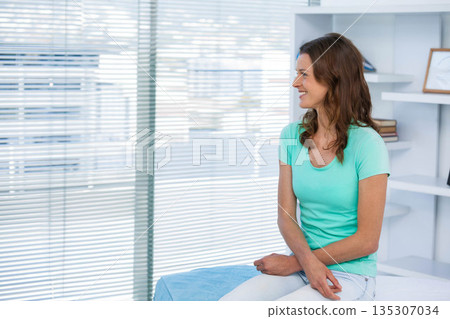 Woman sitting on examination table in exam room with window blinds and framed document, copy space Woman sitting on examination table in exam room with window blinds and framed document, copy space 135307034