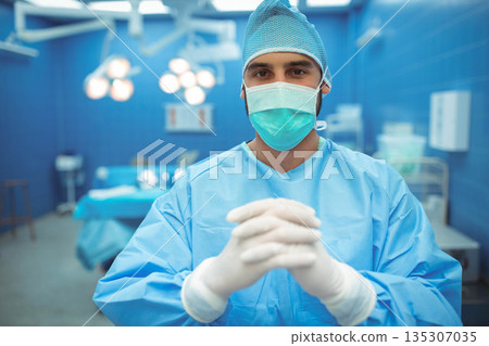Male surgeon standing wearing blue scrubs and mask in operating room with surgical lights 135307035