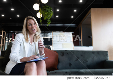 Female business professional holding clipboard while sitting on sofa by whiteboard in office lounge 135307038