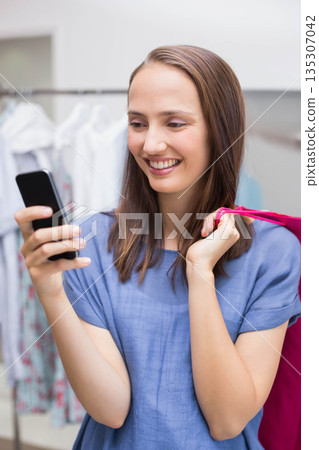 Woman wearing blue blouse holding smartphone carrying pink shopping bag in store with metal racks Woman wearing blue blouse holding smartphone carrying pink shopping bag in store with metal racks 135307042