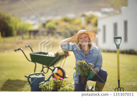 Senior woman kneeling on lawn holding black plastic pot with yellow flowers and adjusting sun hat Senior woman kneeling on lawn holding black plastic pot with yellow flowers and adjusting sun hat 135307051