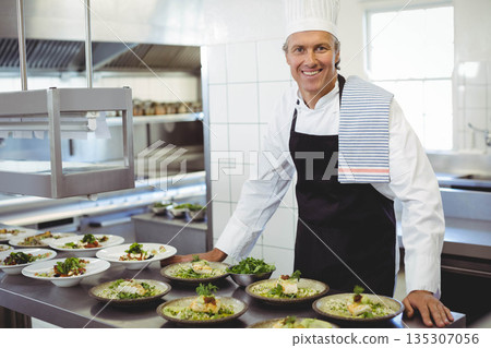 Male chef wearing uniform preparing dishes at table under heat lamps in busy kitchen, copy space Male chef wearing uniform preparing dishes at table under heat lamps in busy kitchen, copy space 135307056