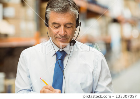 Middle-aged man surveying warehouse aisle wearing shirt and tie with headset and clipboard Middle-aged man surveying warehouse aisle wearing shirt and tie with headset and clipboard 135307057