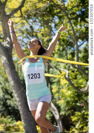 African American woman running and breaking yellow finish line tape across park path with bib 1203 African American woman running and breaking yellow finish line tape across park path with bib 1203 135307058