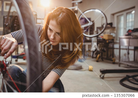 Woman kneeling and adjusting bicycle wheel on repair stand at garage workshop using wrench 135307066
