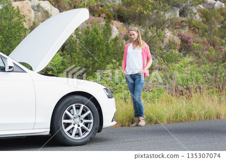 Woman standing on rural roadside beside white sedan with hood open, examining engine compartment 135307074