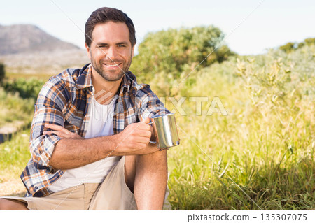 Man sitting on grassy hillside holding stainless steel camping mug while overlooking mountain vista 135307075