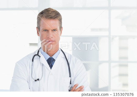 Middle-aged male doctor standing before grid windows in corridor, sporting lab coat and stethoscope Middle-aged male doctor standing before grid windows in corridor, sporting lab coat and stethoscope 135307076