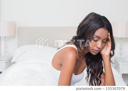 African American woman sitting on bed edge in lit bedroom with pink table lamp, copy space African American woman sitting on bed edge in lit bedroom with pink table lamp, copy space 135307079