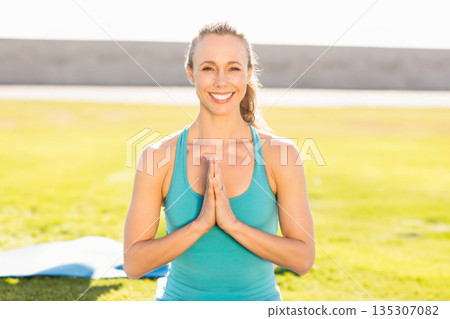 Woman in her 30s kneeling on grassy field beside blue yoga mat, wearing turquoise sportswear 135307082