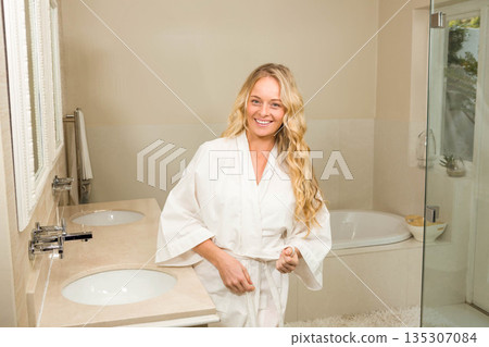 Woman standing in bathroom near sink countertop wearing bathrobe and reaching bath salts by bathtub Woman standing in bathroom near sink countertop wearing bathrobe and reaching bath salts by bathtub 135307084