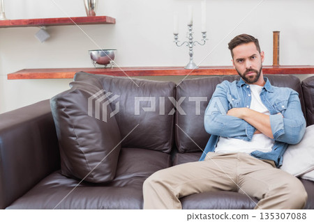 Man sitting on leather sofa with arms crossed in living room beside wooden shelves, copy space 135307088