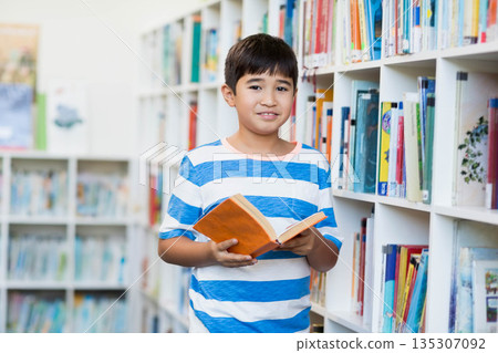 Asian boy reading orange book while standing in library aisle between white bookshelves 135307092