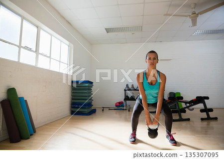 Woman performing kettlebell squat in bright fitness studio with stacked mats and large windows 135307095