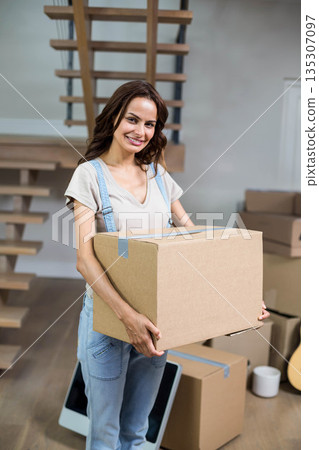 Mid-thirties woman wearing denim overalls holding moving box by monitor and guitar in living room 135307097