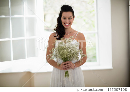 Female bride standing by paned windows holding bouquet in bright suite wearing lace wedding dress 135307108
