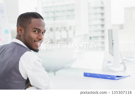 African American man sitting at white desk in office wearing vest viewing monitor and blue binder African American man sitting at white desk in office wearing vest viewing monitor and blue binder 135307116