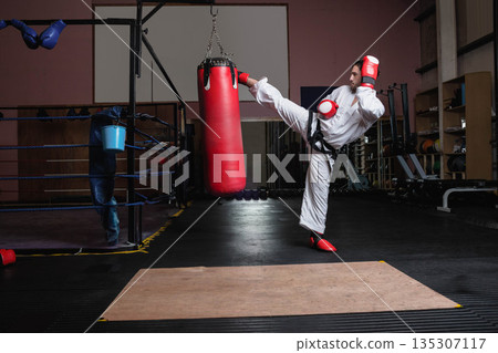 Male karate athlete wearing white uniform delivering side kick to punching bag in dojo, copy space Male karate athlete wearing white uniform delivering side kick to punching bag in dojo, copy space 135307117