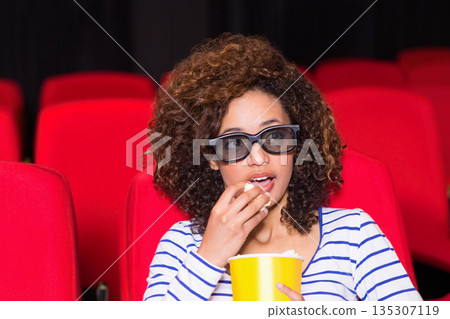 African American woman wearing 3D glasses holding yellow popcorn bucket eating in theater 135307119