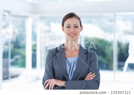 Mid-adult businesswoman standing with arms crossed by office windows overlooking cityscape foliage Mid-adult businesswoman standing with arms crossed by office windows overlooking cityscape foliage 135307121