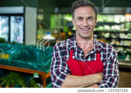 Man standing wearing red apron with arms crossed in produce section among bins, refrigerated cases Man standing wearing red apron with arms crossed in produce section among bins, refrigerated cases 135307140